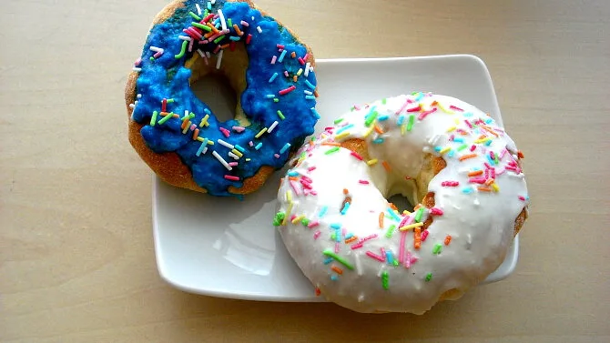 Dva domácí pečené donuty s barevnými cukrovinkami na bílém talíři.
