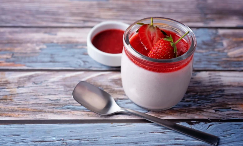 Strawberryová panna cotta v skleněné nádobce s lžící na dřevěném stole.