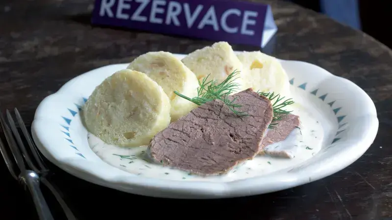 Párek hovězího masa s koprovou omáčkou a knedlíky na talíři.