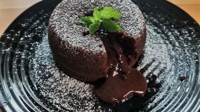 Čokoládový fondant s rozteklou čokoládou na černém talíři posypaný cukrem a ozdobený máty.