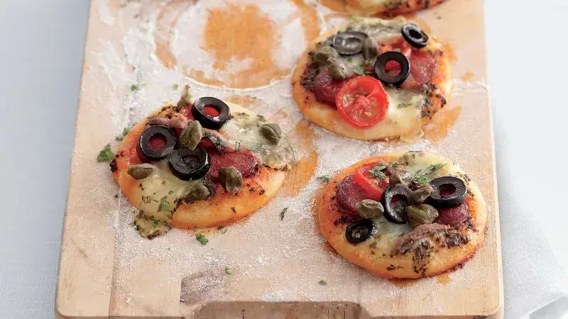 Čtyři malé pizzy s olivami a rajčaty na dřevěném prknu.
