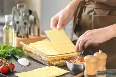 Člověk skládá vrstvy těsta a omáčky do skleněné formy na lasagne.