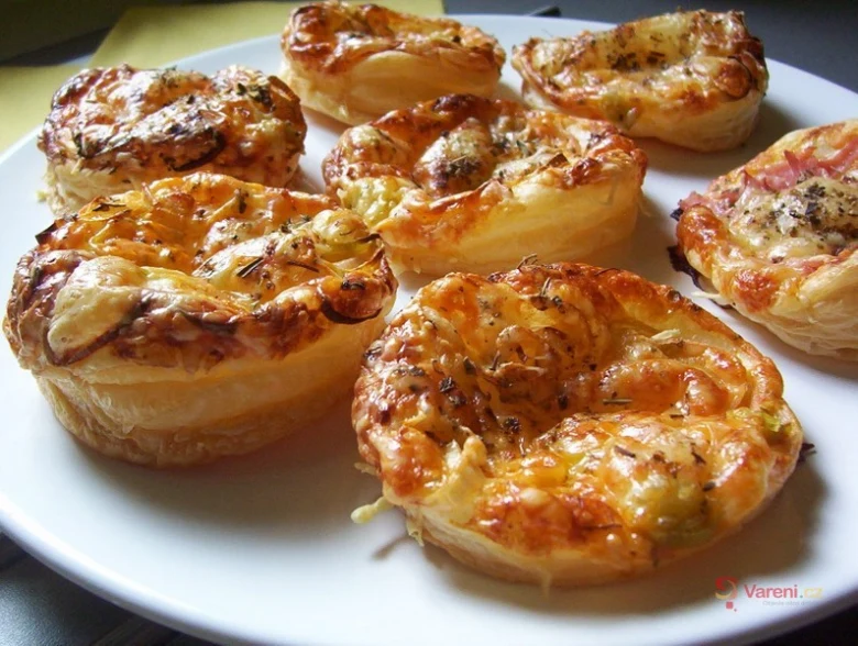Zlatohnědé minipizzy z listového těsta s sýrem a bylinkami na bílém talíři.