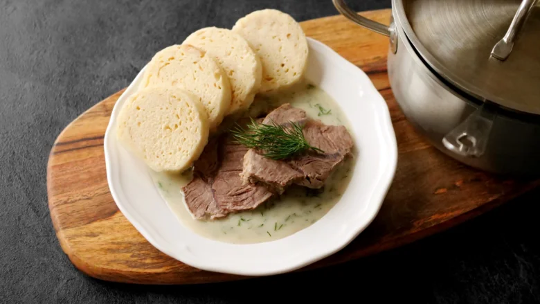Porce hovězího masa s koprovou omáčkou a knedlíky na talíři.