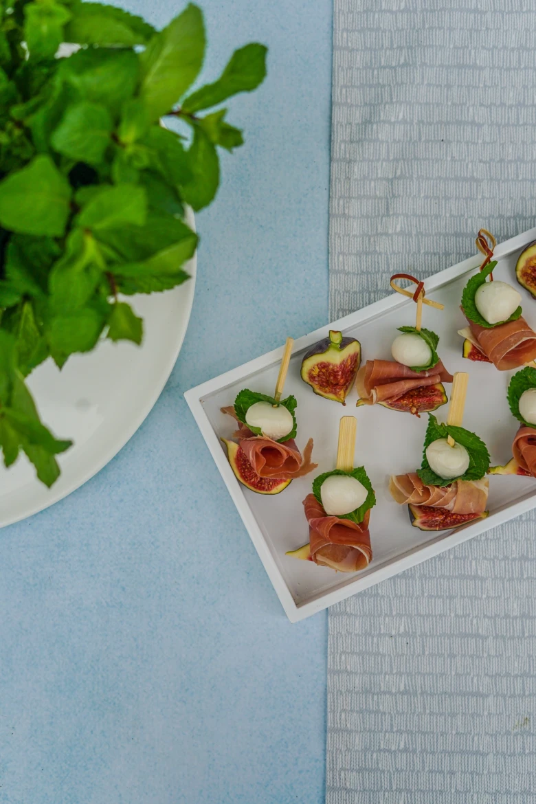 Jednohubky z fíku s prosciutto a mozzarellou na bílém podnose.