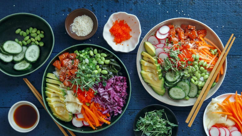 Dva poké bowl s čerstvými zeleninovými ingrediencemi na modrém povrchu.