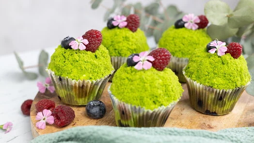 Zelené muffiny s malinami, borůvkami a květy na dřevěném podnose.