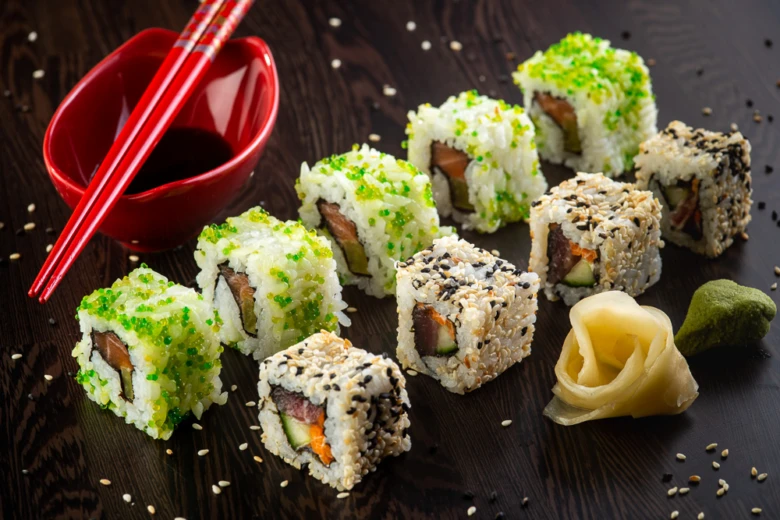 Různé druhy sushi s zelenou i sezamovou omáčkou na tmavém dřevěném podkladu.