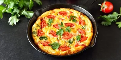 Zeleninová fritáta s rajčaty v železném pánvi na tmavé desce.