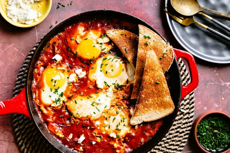 Shakshuka s vejci a toastem v pánvi na červeném podkladu.