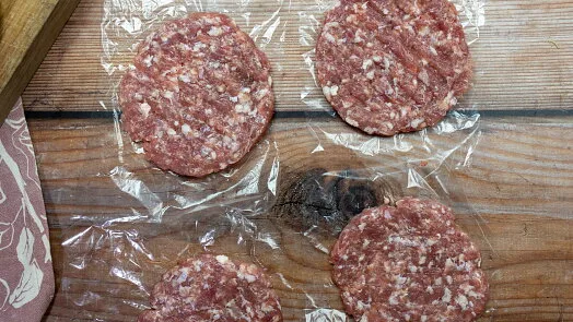 Čtyři syrové hovězí burgerové placky na dřevěném povrchu pod fólií.