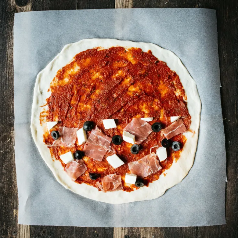 Nepečená pizzy s ingrediencemi na pečicím papíru.