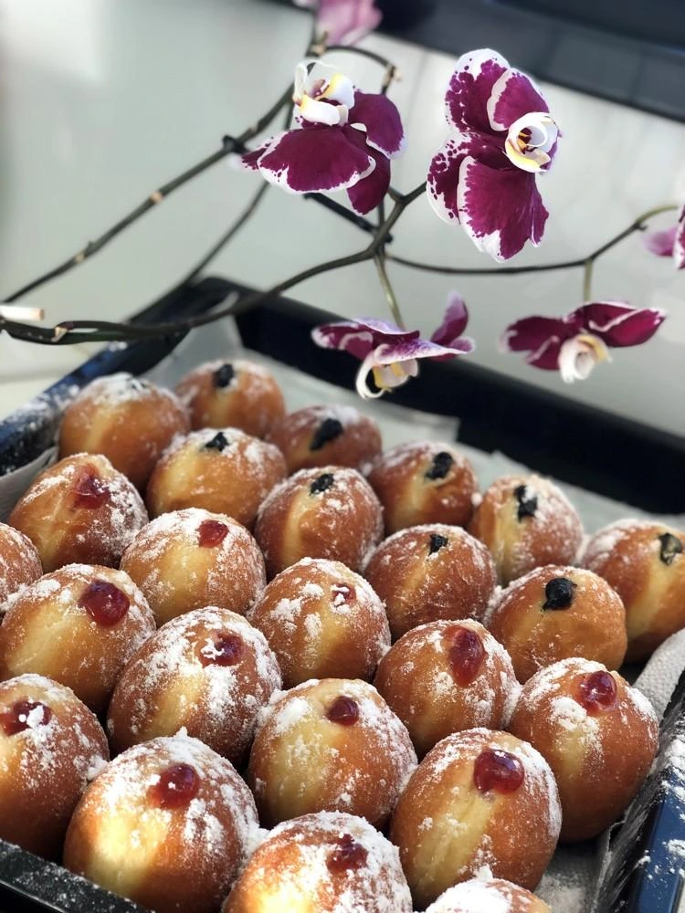Zlatohnědé koblihy posypané moučkovým cukrem s ovocem pod fialovými orchidejemi.