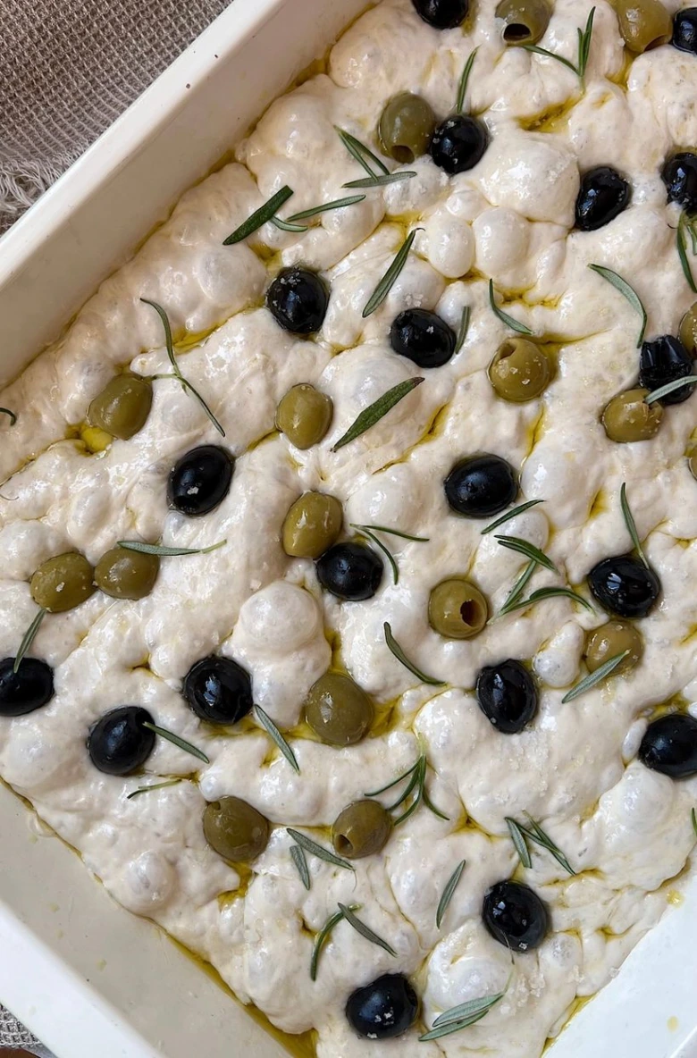 Nepečená focaccia s černými a zelenými olivami v bílé formě.