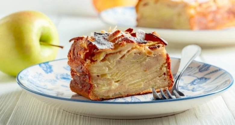 Kousek vrstveného jablečného koláče s moučkou na talíři vedle zelené jablka.