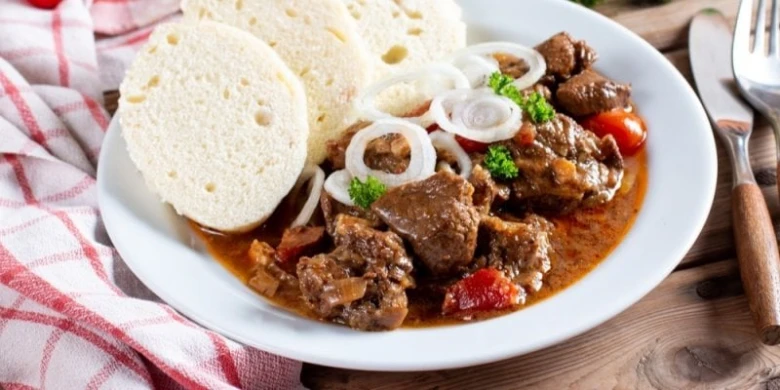 Český guláš s knedlíkem a cibulovými kroužky na talíři.