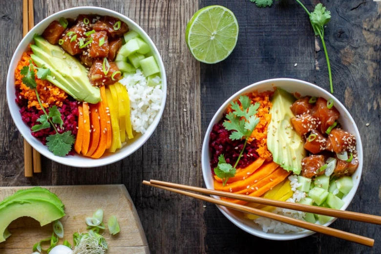 Dva barevné poke bowl s rýží, avokádem, okurkou a rybami na dřevěném stole.