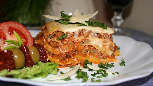 Kousek lasagny s rajčaty a olivami na talíři.