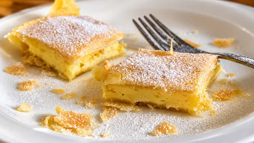 Dva čtvercové kousky tvarohového koláče posypaného cukrem na talíři.